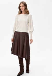 Langärmliges creamfarbenes Oberteil mit Puffärmeln, kombiniert mit einem braunen, kunstledernen Rock mit asymmetrischem Saum und braunen, knielangen Stiefeln.