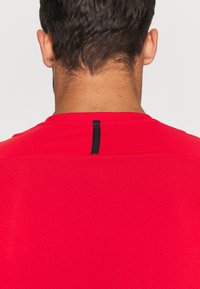 Hombre con cabello corto y oscuro vistiendo una camisa roja perforada con una franja vertical negra en el cuello de la parte posterior, frente a un fondo blanco.