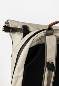 Rucksack mit Rollverschluss in beigeem Außenmaterial, schwarzem Innenfutter, verstellbaren Trägern und Schnallenverschluss. Strukturierte Oberfläche mit elegantem Design.