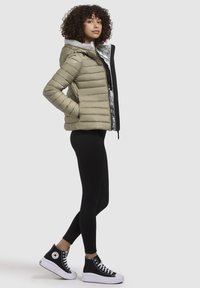 Licht olijfgroene pufferjack met een zilverkleurige capuchon, gecombineerd met zwarte leggings en zwarte high-top sneakers met witte zolen.