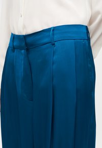 Μπλε σατέν παντελόνι με ψηλή μέση και θηλιές για ζώνη, συνδυασμένο με λευκό πουκάμισο κουμπωτό που έχει μπει μέσα στο παντελόνι, εμφανίζοντας την πλαϊνή τσέπη και τις πιέτες στο μπροστινό μέρος.