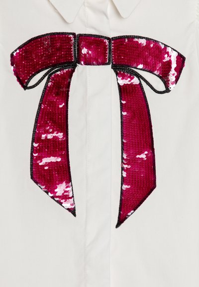 Vit skjorta med en stor applikation av paljetter i fuchsia formad som en rosett med svart kontur och detaljer i mitten.