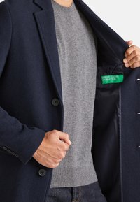 Abrigo de lana azul marino con solapa muesca, cierre de botón único y suéter de punto gris debajo. La etiqueta interior presenta "United Colors of Benetton."