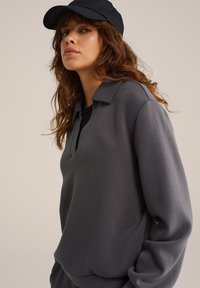 Mujer con cabello largo y castaño que lleva una gorra negra y una camisa de manga larga en gris oscuro con cuello, mirando ligeramente de lado contra un fondo simple.