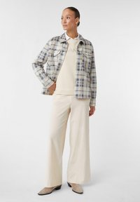Giacca camicia a quadri, blu chiaro e crema, con due tasche sul petto, indossata sopra un maglione beige. Abbinata a pantaloni larghi crema e scarpe beige.