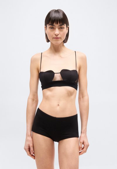 Mujer con cabello corto y oscuro, vestida con un bralette negro de tirantes finos y braguitas a juego de talle alto, de pie frente a un fondo claro y sencillo.