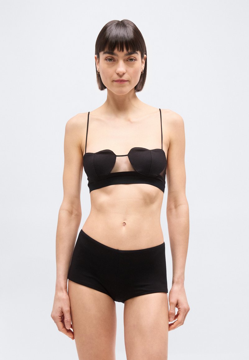 Γυναίκα με κοντά σκούρα μαλλιά, φοράει μαύρο τοπ με λεπτές τιράντες και ταιριαστό σλιπ με ψηλή μέση, στέκεται μπροστά σε απλό φωτεινό φόντο.