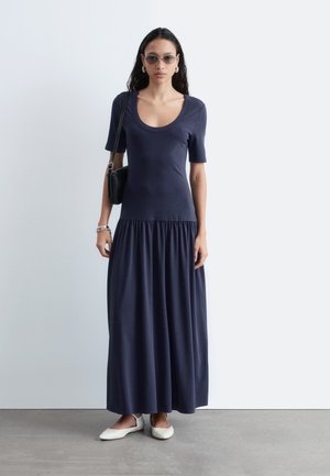 Vrouw staand, gekleed in een marineblauwe maxi-jurk met korte mouwen en een ronde hals, witte platte schoenen, zonnebril, oorringen en een zwarte schoudertas dragend.