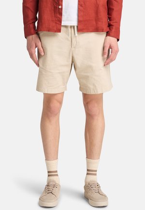 Shorts de lino beige con cintura elástica y detalle de cordón, combinados con zapatillas beige y calcetines de canalé crema con rayas marrones.