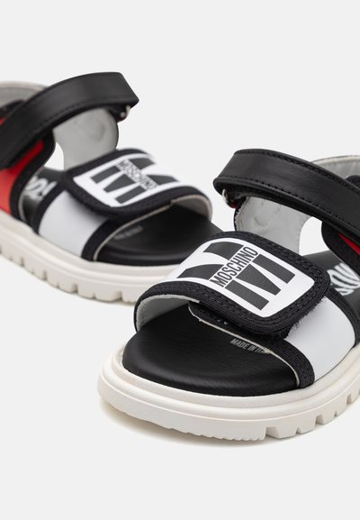 Sandalias en blanco y negro con correas de velcro, suela de goma y un acento rojo. Presentan un patrón de logotipos en las correas y una plantilla texturizada.