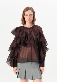 Vrouw in sheer donkerbruin blouse met grote ruffles, gecombineerd met een knielange lichtgrijze plissérok, staande tegen een effen witte achtergrond.