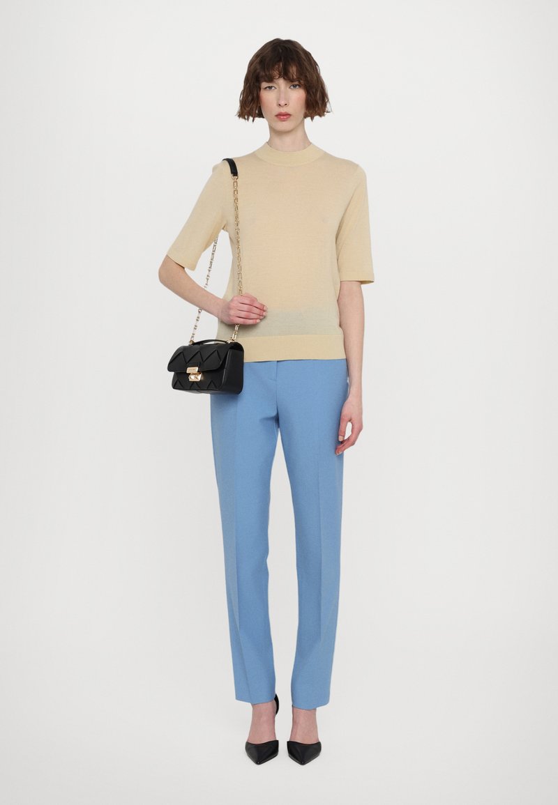 Beige gestricktes Kurzarmoberteil, hellblaue maßgeschneiderte Hose und eine kleine schwarze Handtasche mit Kettenriemen und goldenen Beschlägen; schwarze spitze High Heels.