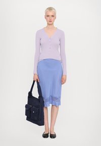 Lichtpaarse blouse met lange mouwen en knopen, blauwe rok tot de knie met een doorschijnende zoom, en een donkerblauwe tas met twee voorzakken. Platte zwarte schoenen.