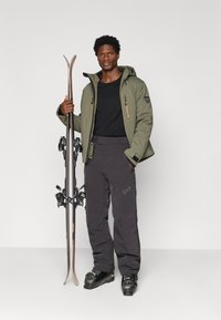 Síruha olíva zöld dzsekivel és sötétszürke nadrággal, cipzárakkal és logókkal díszítve, sílécekkel és csizmákkal kiegészítve.