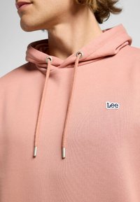 Felpa rosa chiaro con cappuccio regolabile, dettagli in metallo argentato e un piccolo logo ricamato "Lee" sul petto. Tessuto morbido e strutturato.