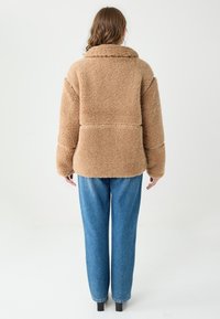 Brązowa, sztuczna futrzana oversize kurtka z kołnierzem, z poziomym przeszyciem na plecach i przodzie, w połączeniu z jasnoniebieskimi szerokimi spodniami jeansowymi.