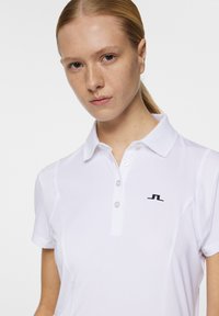 Valkoinen polo-paita, jossa on kaulus, kolme nappia ja musta logo rinnassa. Liukas pinta, jossa on hienovaraisia suunnitteluyksityiskohtia hartioilla.
