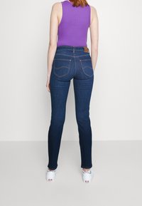 Vaqueros ajustados azul oscuro con bolsillos traseros y costuras contrastantes, combinados con una camiseta sin mangas morada y zapatillas blancas.