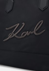 Musta kangaskassi, jossa on keskeinen, tyylitelty harmaa brodeerattu signeeraus. Sileä pinta, jossa on ommeltuja yksityiskohtia.