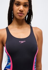 Νεαρή γυναίκα με πλεγμένα μαλλιά, φοράει μαύρο μαγιό Speedo με ροζ και μπλε πλευρικές λεπτομέρειες, κοιτάζοντας προς τα εμπρός μπροστά σε απλό φόντο.