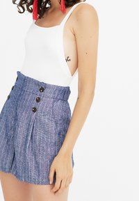 Un body blanc à bretelles larges associé à un short bleu rayé taille haute avec une fermeture à bouton et des plis sur les côtés.