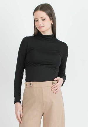 Vrouw met lang bruin haar die een zwarte coltrui met lange mouwen en beige hooggesneden broek draagt tegen een effen achtergrond.