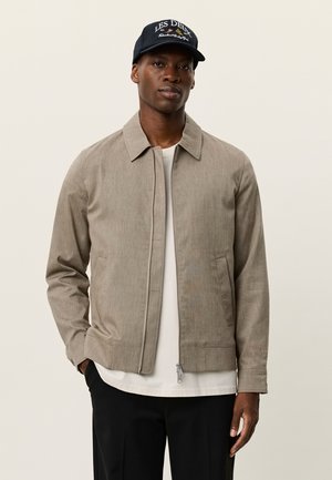 Chaqueta marrón claro con textura, cierre frontal con cremallera, cuello puntiagudo y bolsillos laterales. Se lleva sobre una camisa blanca, combinada con pantalones negros y una gorra.