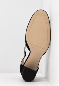 Chaussure à talon haut en velours noir avec une semelle beige lisse, présentant des côtés découpés et un petit talon. Logo de marque notable au dos.