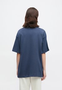 Άτομο με καστανά μαλλιά μέχρι τους ώμους, φορώντας ένα χαλαρό σκούρο μπλε κοντομάνικο μπλουζάκι και ανοιχτόχρωμα παντελόνια, γυρισμένο μακριά σε λευκό φόντο.