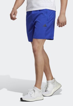 Mężczyzna w niebieskich sportowych szortach z logo Adidas i białych butach do biegania, stojący z jedną nogą lekko wysuniętą do przodu.