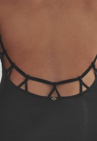 Schwarzer Badeanzug mit einem Riemenrücken-Design, das verflochtene Träger und eine kleine goldene Hardware-Details in der Mitte aufweist.
