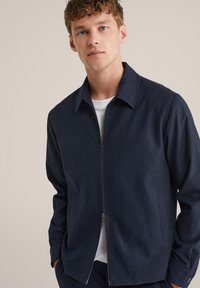 Temno modra zip-up jakna iz teksturiranega materiala, z ovratnikom, navpičnimi črtami in stranskimi žepi.
