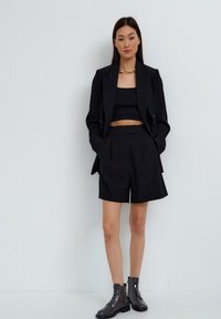 Zwarte getailleerde blazer over een zwarte crop top en bijpassende high-waisted shorts. Afgemaakt met zwarte glanzende enkellaarsjes en een.chunky gouden ketting.
