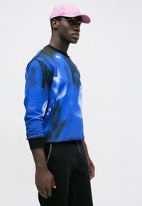 Hombre con una gorra rosa, sudadera azul con un estampado y puños negros, y pantalones negros, de pie contra un fondo blanco liso.