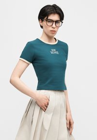 Νέα γυναίκα με στρογγυλά γυαλιά, μπλε-πράσινο t-shirt New Balance και εκρού πλισέ φούστα, με το χέρι στην τσέπη, στέκεται μπροστά σε λευκό φόντο.