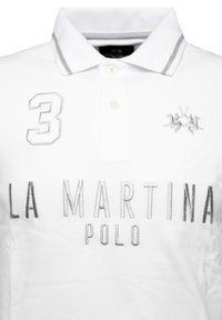 Vit polo-shirt med grå detaljer, som har ett broderat nummer "3" och "LA MARTINA POLO" i silver över framsidan.