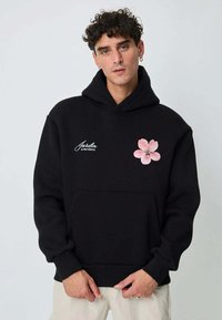 Homme aux cheveux courts et bouclés portant un sweat à capuche noir avec une fleur rose et le texte « Jardin de Provence », mains jointes, pantalon clair.