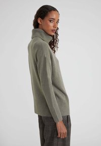 Donna che indossa un maglione girocollo verde oliva largo e pantaloni a quadri grigi, in profilo contro uno sfondo chiaro e semplice.