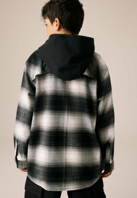 Chemise à carreaux noir et blanc avec une capuche noire, présentant une coupe décontractée, des poignets à boutons et une texture de tissu douce.