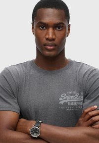 Grå kortærmet t-shirt med "SuperDry" logo i hvid, båret med et sort læderrem ur med rund urskive og sølvdetaljer.
