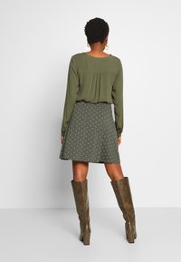 Blouse à manches longues vert olive avec un dos plissé, associée à une jupe longueur genou à motif à pois blancs et des bottes marron texturées jusqu'aux genoux.