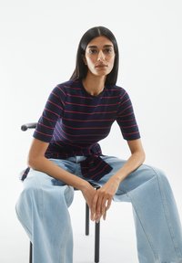 Navy and red striped short-sleeve top with a tie detail at the waist, paired with light blue wide-leg jeans, seated on a black chair.