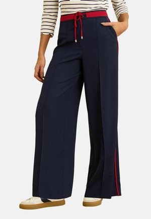 Marineblauwe broek met wijde pijpen, rode tailleband en zijstreep, gedragen met een gestreept shirt en witte sneakers met bruine zolen.