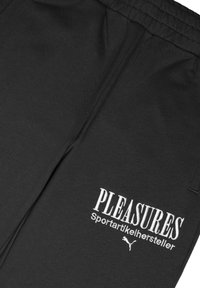 Schwarze Sportpants aus glattem Stoff, mit "PLEASURES" und "Sportartikelhersteller" in Weiß gestickt, versehen mit einem Puma-Logo.