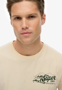 Camiseta de cuello redondo beige hecha de algodón. Presenta un gráfico de una montaña verde y un árbol en el lado izquierdo del pecho. Textura suave, corte casual.