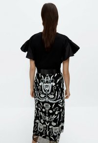 Robe noire avec des manches bouffantes transparentes, ornée d'une broderie en argent complexe, au design fluide et à une ceinture noire ajustée à la taille.