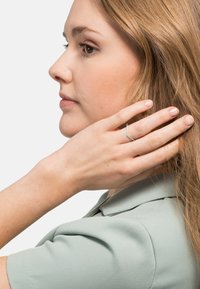 Silberner Ring mit Zickzack-Design, der kleine eingearbeitete Steine aufweist, getragen an einer Hand in der Nähe des Gesichts. Texturiertes hellgrünes Blusentop ist sichtbar.