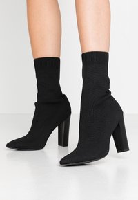 Czarne botki na wysokim obcasie do kostki z dzianinową cholewką w stylu skarpety i spiczastym noskiem. Posiadają masywny obcas oraz teksturowane elastyczne wykończenie na górze.