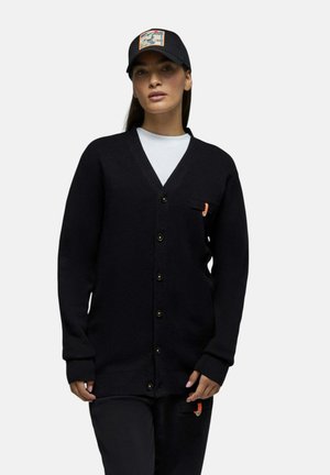 Femme portant un cardigan noir boutonné, un pantalon noir, une chemise blanche et une casquette de baseball noire avec un patch coloré, debout devant un fond uni.