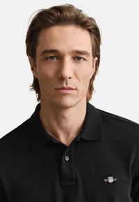 Juoda polo marškinėlių su trimis mygtukais, trumpomis rankovėmis ir siuvinėta krūtinės logotipu. Slystanti tekstūra ir klasikinis kirpimas.